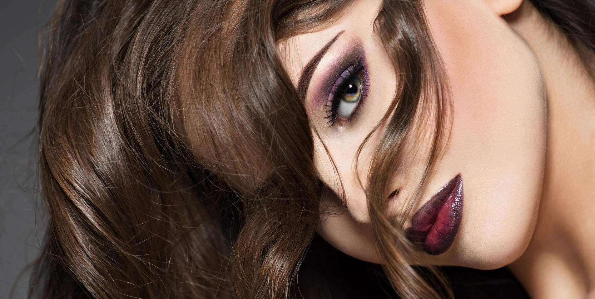 Verführerischer Blick Eine junge Frau mit langem, braunem Haar, violettem Lidschatten und dunkelrotem Lippenstift hält ihren Kopf schief.