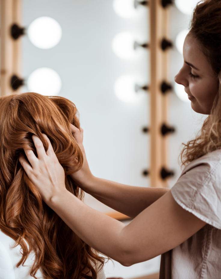 Perfektes Haarstyling Eine junge Stylistin in einer beigen Bluse knetet die langen, roten Locken einer Kundin.